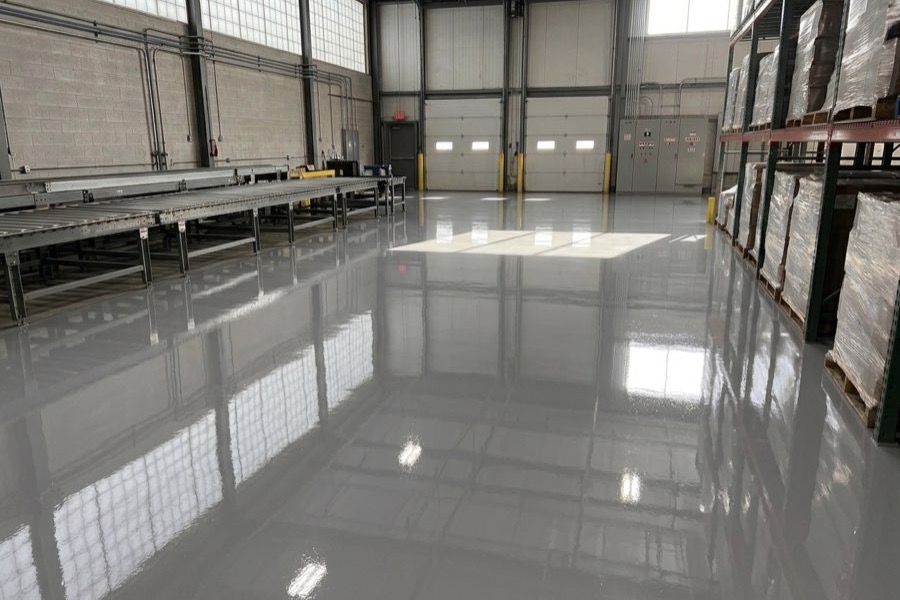 A warehouse floor in a grey high-gloss coating, running between a conveyor line and loaded pallet racking toward the loading doors.